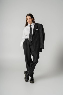 full length of young woman in elegant formal wear and rough leather boots posing with hand in pocket isolated on grey