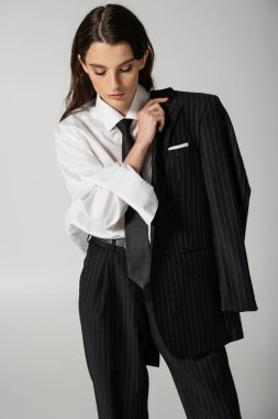 young and trendy woman in elegant formal wear holding black striped blazer isolated on grey