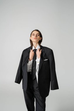 brunette woman in formal wear holding black oversize blazer and looking away isolated on grey clipart