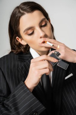 young and stylish woman with makeup lighting cigarette isolated on grey