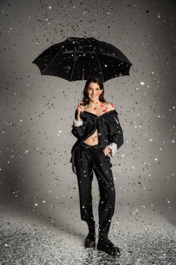 happy young woman in stylish clothes standing with black umbrella under shiny confetti on grey background