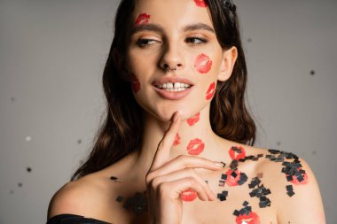 sensual woman with red lipstick marks on face and confetti on bare shoulders touching chin and looking away on grey  clipart