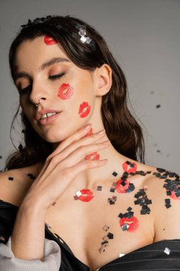 sensual woman with red kiss prints and shiny confetti on face and naked shoulders posing with closed eyes on grey 