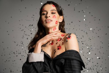 brunette woman in black blazer with bare shoulders and lip prints on body standing under shiny confetti on grey background