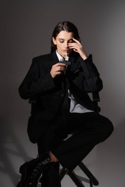fashionable woman in black pantsuit lighting cigarette while sitting on dark grey 