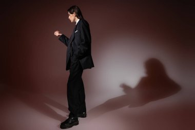 side view of young woman in elegant oversize pantsuit and rough boots standing on brown background