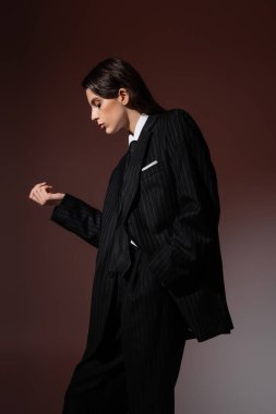 side view of brunette woman in black striped blazer posing with hand in pocket on brown background