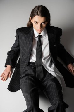 high angle view of fashionable model in oversize shirt and black pantsuit sitting on grey background