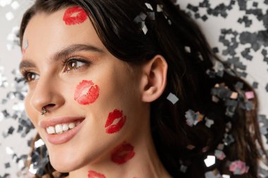 portrait of pleased woman with piercing and red kiss prints looking away near silver confetti on grey background