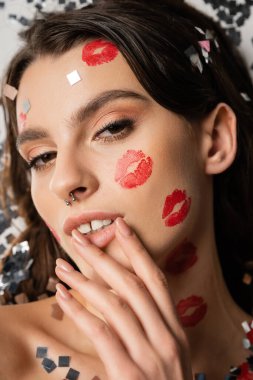 portrait of sensual woman with makeup and red kiss prints touching lips and looking at camera near silver confetti on grey 