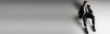 high angle view of stylish woman in formal wear and laced-up boots sitting on grey background, banner