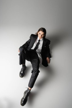 high angle view of young woman in elegant formal wear and rough boots looking at camera while sitting on grey 