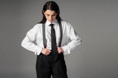 brunette woman in white shirt and black tie adjusting oversize trousers with suspenders isolated on grey