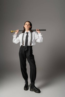 full length of young model in elegant attire and rough leather boots posing with walking cane on grey background