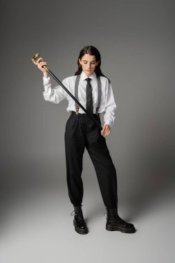 full length of stylish woman in elegant oversize outfit and laced-up boots holding walking cane on grey 