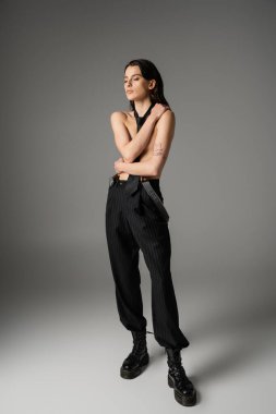 full length of sexy shirtless woman in trendy pants and rough leather boots hiding bust while standing on grey background