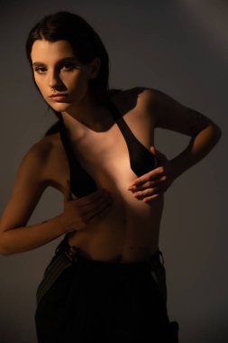 young shirtless woman in black pants and breast tape looking at camera on grey background with lighting