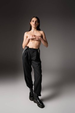 shirtless woman with wet body covering breast with hands while posing in black pants and rough boots on grey 