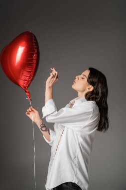 side view of young tattooed woman in white shirt holding cigarette near red festive balloon on grey 