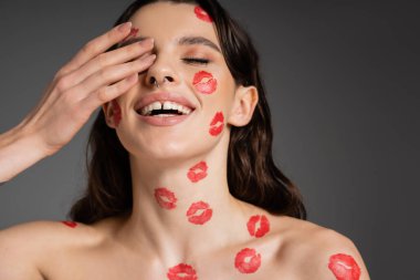 cheerful woman with red lip prints on face and bare shoulders covering eye with hand isolated on grey