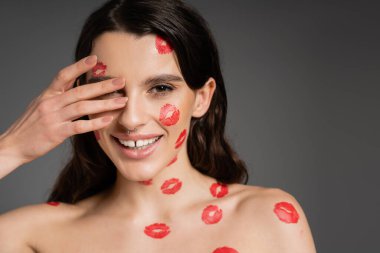 sexy brunette woman with red lipstick marks on bare shoulders and face covering eye with hand and smiling isolated on grey
