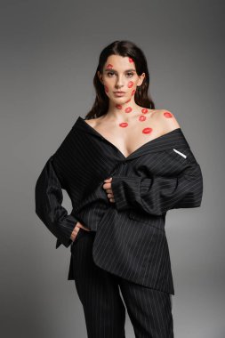 sexy woman with red lip prints on face and naked shoulders posing in black oversize pantsuit isolated on grey