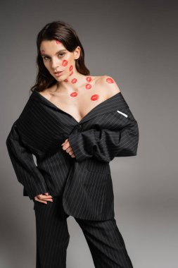 seductive woman in black oversize suit posing with red lip prints on face and bare shoulders isolated on grey