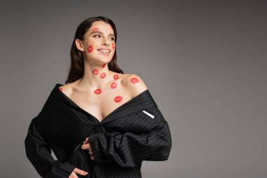 cheerful young woman with red kisses on face and body looking away while posing in oversize blazer isolated on grey