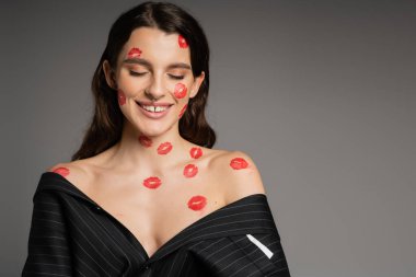 happy young woman with red lipstick marks on face and bare shoulders smiling with closed eyes isolated on grey