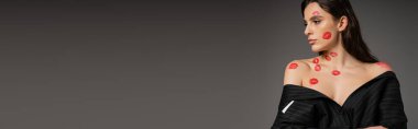 pretty woman with red lipstick marks on naked shoulders posing in black oversize blazer isolated on grey, banner