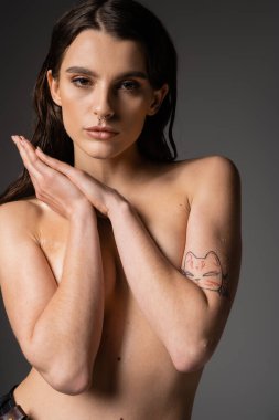 sexy tattooed woman with shirtless body hiding breast with arms and looking at camera on grey background