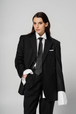 young and fashionable woman in black oversize pantsuit holding hand in pocket while looking at camera isolated on grey