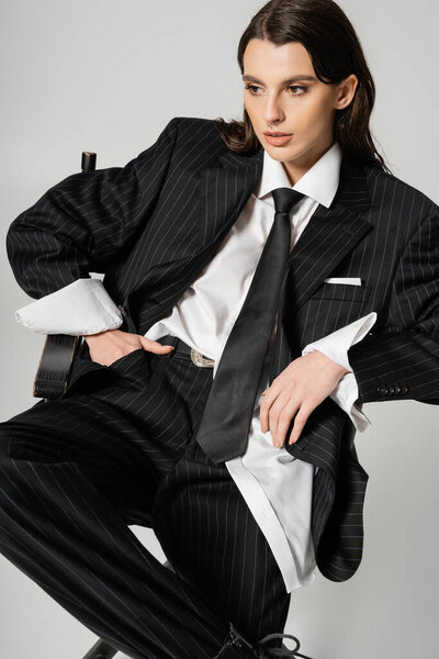 trendy woman in black oversize suit and white shirt holding hand in pocket while sitting isolated on grey