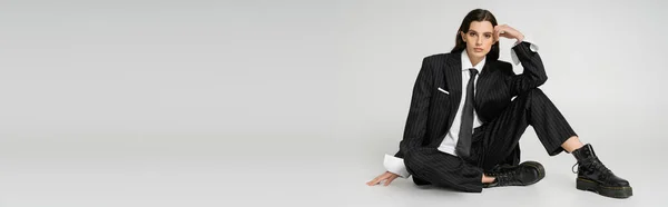 stock image stylish woman in elegant formal wear and rough laced-up boots holding hand near head while sitting on grey background, banner