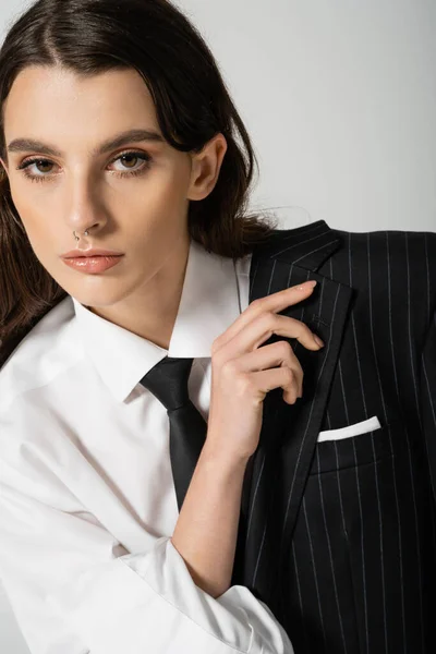 stock image portrait of young woman in white shirt and tie holding black blazer and looking at camera isolated on grey