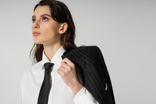 brunette woman in white shirt and tie holding black striped blazer while looking away isolated on grey