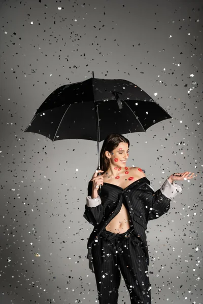 happy woman with red kiss prints on body wearing trendy clothes and standing under black umbrella and confetti on grey