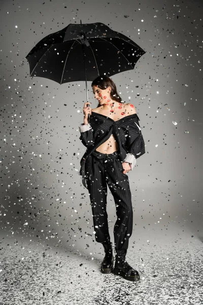 stock image happy woman in black stylish clothes standing with umbrella under sparkling confetti on grey 