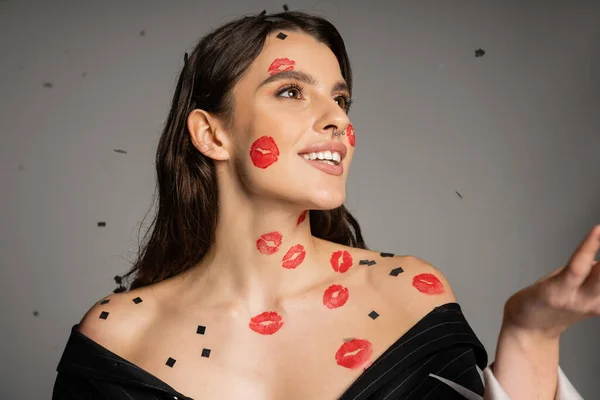 cheerful woman with red lip prints and confetti on face and bare shoulders looking away on grey background