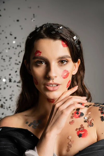 charming woman with red kiss prints on face looking at camera near sparkling confetti on grey