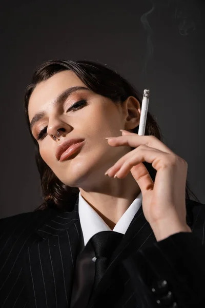 stock image portrait of young brunette woman in elegant attire holding cigarette and looking at camera isolated on dark grey