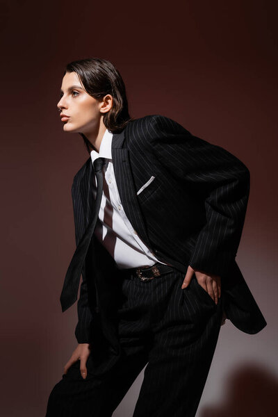 fashionable woman in black and striped suit and white shirt with tie looking away while posing on brown 