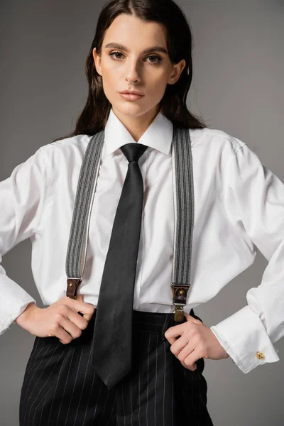brunette woman in white oversize shirt and tie holding black pants with suspenders and looking at camera isolated on grey