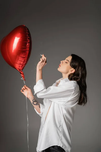 stock image side view of young tattooed woman in white shirt holding cigarette near red festive balloon on grey 