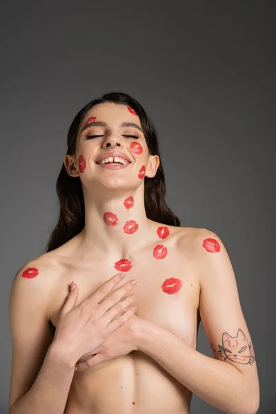 joyful shirtless woman with red kisses on face and body covering breast with hands isolated on grey