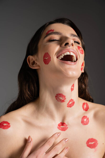 возбужденная женщина с закрытыми глазами и красными поцелуями на лице и обнаженными плечами смеется изолированно на сером