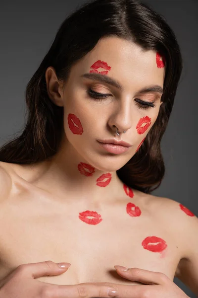 sensual shirtless woman with red kisses on body and face covering breast with hands isolated on grey