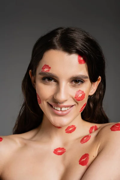 young and happy woman with red kiss prints on face and bare shoulders looking at camera isolated on grey
