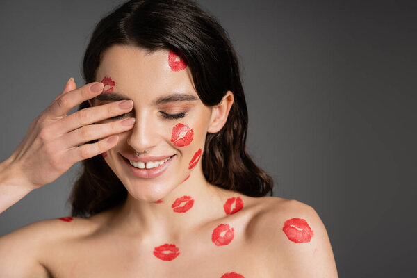 улыбающаяся женщина с красными отпечатками поцелуев на теле и лице, закрывая глаза рукой, изолированной на сером