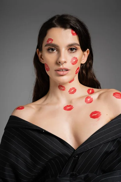 young brunette woman with lip prints on face and bare shoulders looking at camera isolated on grey